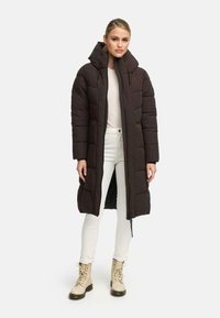 Lång, mörkbrun puffjacka med hög krage, dragkedjestängning och sidofickor. Stylad med ljusa jeans och beige snörstövlar.
