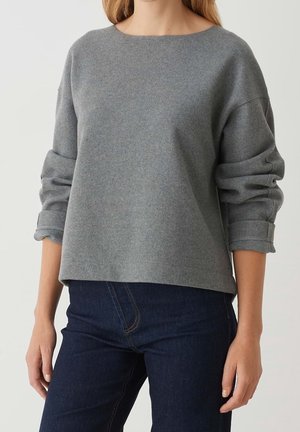 Femme portant un pull gris ample à manches longues retroussées et un jean bleu foncé, vue du cou jusqu'aux hanches.