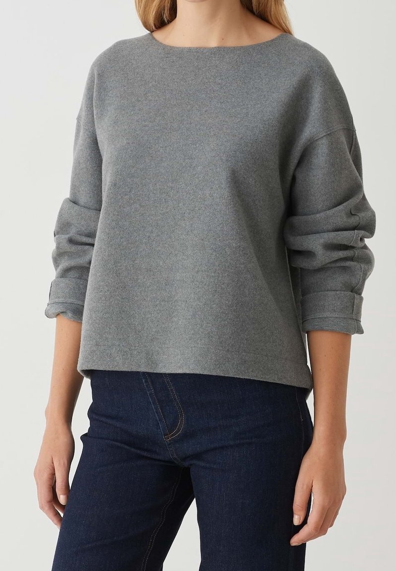 Femme portant un pull gris ample à manches longues retroussées et un jean bleu foncé, vue du cou jusqu'aux hanches.