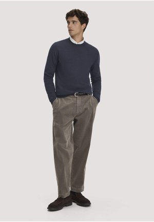 Junger Mann, der einen marineblauen Pullover, ein weißes Hemd mit Kragen, beige Cordhosen, einen schwarzen Gürtel und dunkelbraune Schuhe trägt, steht mit den Händen in den Taschen.
