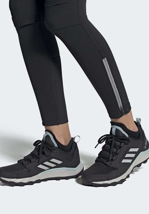 Scarpe da ginnastica nere con strisce bianche indossate con leggings neri dotati di cerniere riflettenti alla caviglia su uno sfondo grigio chiaro.