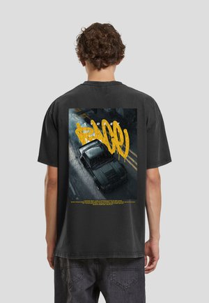Joven con camiseta negra que muestra un gráfico de un coche negro derrapando en una calle con texto amarillo al estilo graffiti.