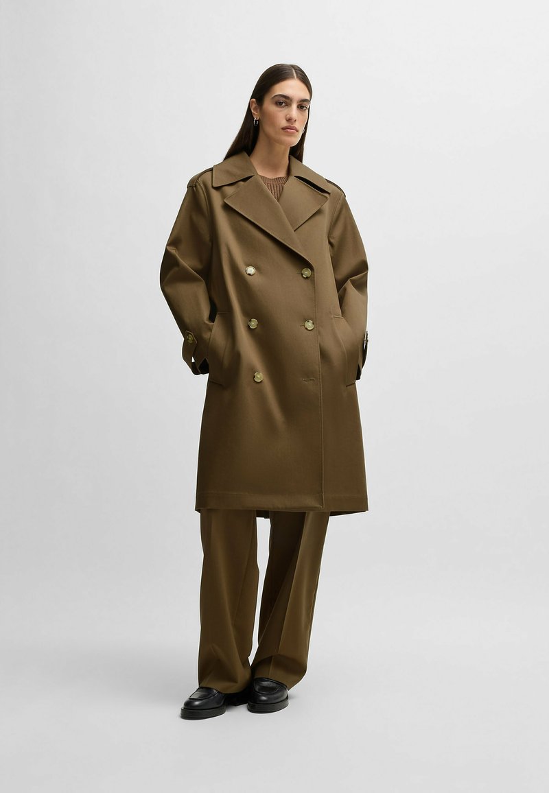 Cappotto trench doppiopetto di colore khaki, realizzato in tessuto spesso. Presenta ampie lunette, bottoni dorati e tasche laterali. Abbinato a pantaloni a gamba larga.