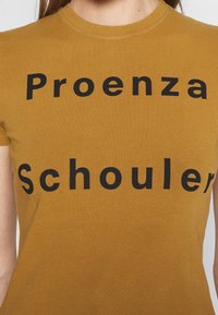 Senapsgul bomulls t-shirt med rund halsringning och fet svart text som står "Proenza Schouler" tvärs över framsidan i versaler.