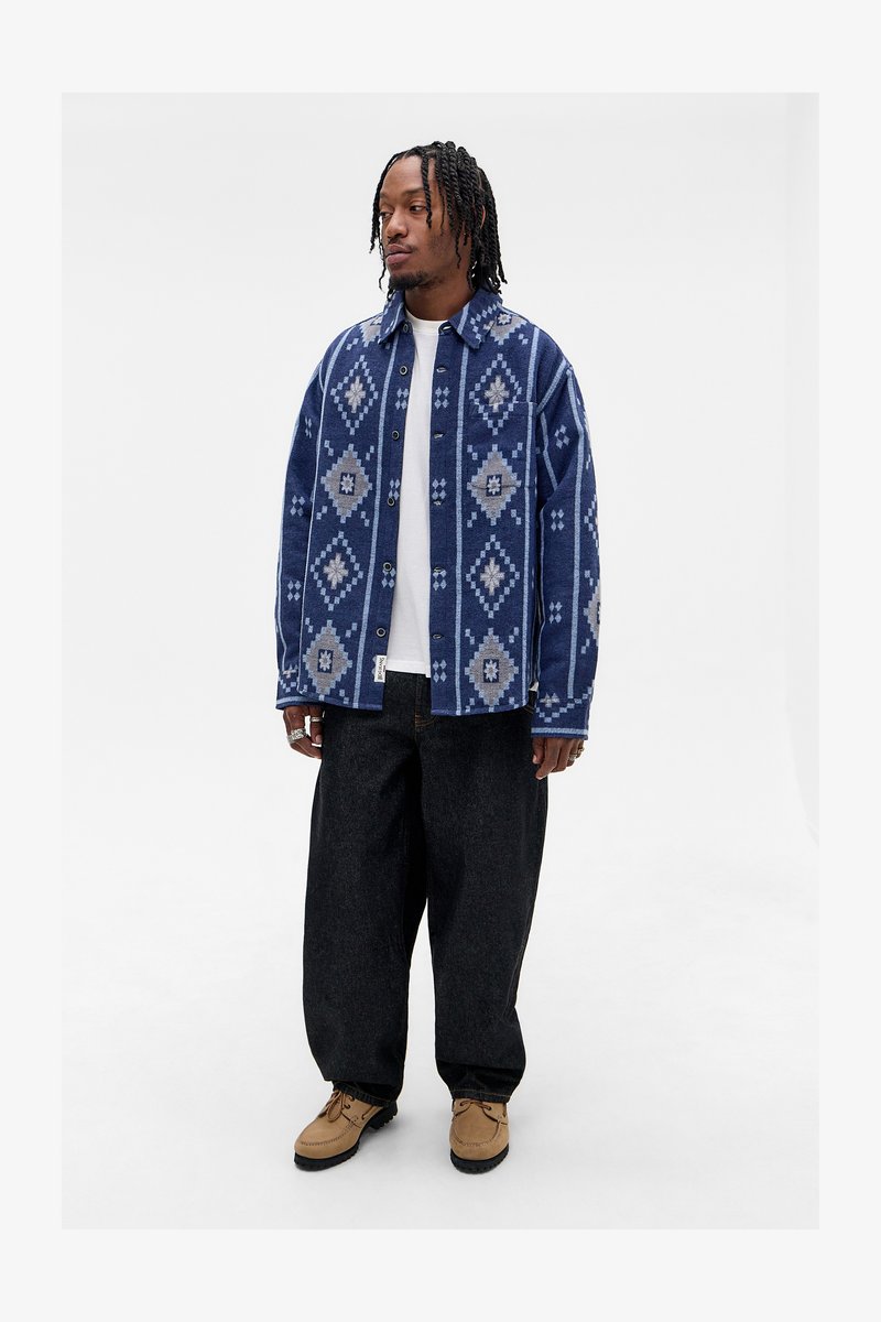 Homemem em pé, vestindo uma camisa de botão azul com padrão sobre uma t-shirt branca, calças largas pretas, sapatos castanhos e vários anéis, contra um fundo liso.