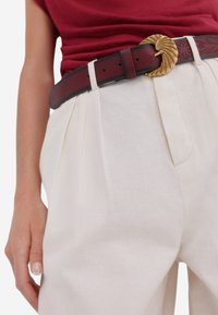 Bordeaux leren riem met een gouden, spiraalvormige gesp, gedragen met crèmekleurige, geplooide linnen broek. Zachte textuur en gepolijste afwerking.