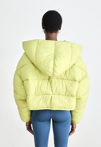 Puffet, kort jakke i lys limegrøn, med hætte, quiltet design og tekstureret stof. Bagfra ses den ovale form.