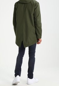 Olivově zelený dlouhý kabát s kapucí, hladkým povrchem, prodlouženým zadním lemem, kombinovaný s tmavými džínami a bílými teniskami. Jednoduchý design.