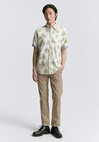 Chemise à manches courtes boutonnée crème avec un motif de feuilles de palmier, associée à un pantalon beige et des chaussures en cuir noires. Accents des poignets bleu clair.