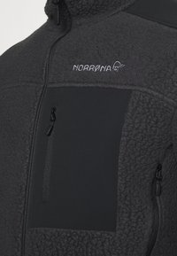 Giacca in pile nera con un pannello liscio, tasca petto con zip e logo ricamato. Tessuto testurizzato sul corpo per il calore.