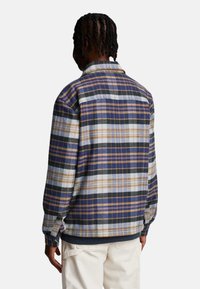 Camisa de franela a cuadros para hombre que presenta una mezcla de colores azul, negro y beige, con un corte relajado, cuello de solapa y puños abotonados.