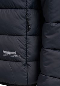 Donkerblauw gewatteerd jack met doorgestikt ontwerp, zwarte ritssluiting en logobeschermer. Gladde, glanzende stof met een lichtgewicht, geïsoleerde uitstraling.