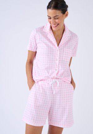 Donna che indossa una camicia a maniche corte con bottoni a quadri rosa e bianchi e pantaloncini con coulisse abbinati, sorridente con le mani nelle tasche.