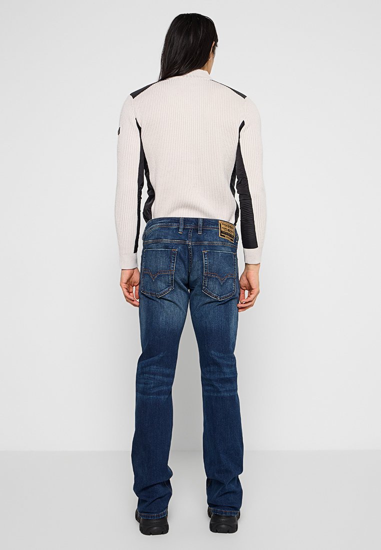 Rippiger cremefarbener Pullover mit schwarzen Schultereinsätzen, kombiniert mit dunkelblauen Jeans aus Denim mit Patches und zwei hinten liegenden Taschen. Schwarze Schuhe sind sichtbar.