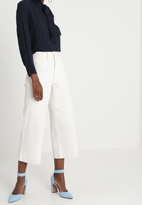 Henkilö, joka on pukeutunut tummansiniseen pitkähihaiseen paitaan, jossa on solmio, ja valkoisiin leveisiin kaprihousuihin, jotka on yhdistetty vaaleansinisiin nilkkaremmiin kenkiin.