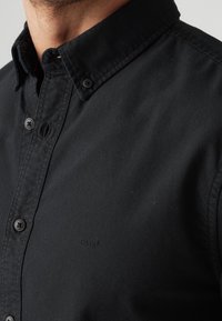 Schwarzes Hemd zum zuknöpfen mit Kragen, aus glattem Stoff; verfügt über schwarze Knöpfe und ein kleines, dezentes Logo auf der Brust.