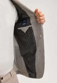 Strukturierter brauner und weißer Blazer mit einem gemusterten Innenfutter. Verfügt über eine schlanke Passform, eine Innentasche und dezente graue Akzente.