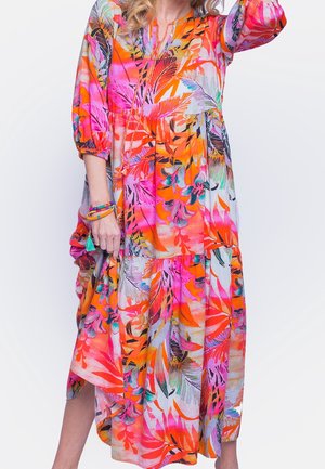 Kvinde iført en lys, flerfarvet blomsterkjole med orange, pink og lilla toner, holder kjolens skørt med den ene hånd og bærer perlearmbånd.