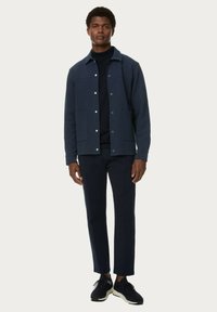 Giacca blu navy con bottoni e due tasche frontali, indossata sopra un dolcevita nero, abbinata a pantaloni slim-fit coordinati e sneakers scure.