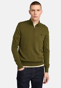 Olivgrüner Pullover mit halber Reißverschluss-Kragen, gerippten Bündchen und Saum. Baumwollmaterial mit dezentem Logodetail auf der Brust. Dunkelblaue Jeans getragen.