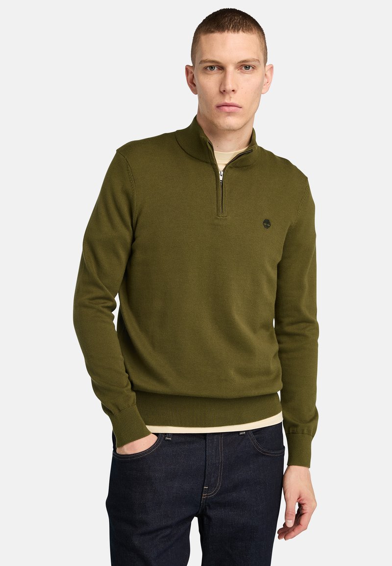 Olivgrüner Pullover mit halber Reißverschluss-Kragen, gerippten Bündchen und Saum. Baumwollmaterial mit dezentem Logodetail auf der Brust. Dunkelblaue Jeans getragen.