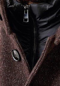 Cappotto marrone caratterizzato da un esterno in lana testurizzata, una morbida fodera interna nera, grande bottone e chiusura con zip con dettagli in metallo.