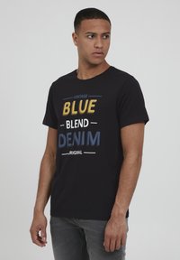 Melna kokvilnas T-krekls ar izteiksmīgu tekstu dzeltenā, baltā un zilā krāsā, kas lasāms "VINTAGE BLUE BLEND DENIM ORIGINAL." Īsas piedurknes, apaļa kakla izgriezums.