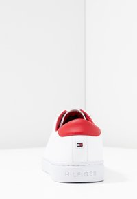 Zapatilla blanca con una pestaña de talón de cuero rojo, textura lisa y detalle sutil del logo. Cuenta con un cuello acolchado y suela de goma.