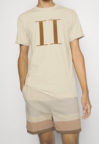 Hombre vistiendo una camiseta beige con un gran gráfico marrón de "II" y pantalones cortos a rayas beige y marrones, de pie frente a un fondo blanco liso.