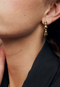 Guldtonade örhängen med tre runda pärlor i graderade storlekar, med en slät och glansig yta, bärs tillsammans med mörka kläder.