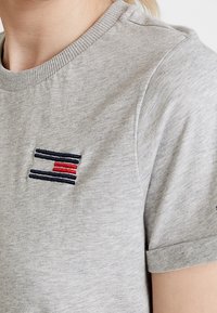 Šedé bavlněné tričko s krátkým rukávem, s malým vyšívaným logem ve navy a červených pruzích na hrudi. Měkká textilie.