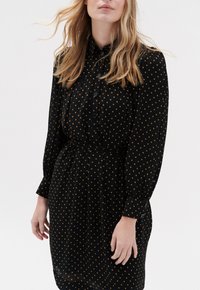 Zwarte jurk met lange mouwen en knoopmanchetten, gekleed met gele stippen en een riem op de taille, wat een getailleerde uitstraling geeft.