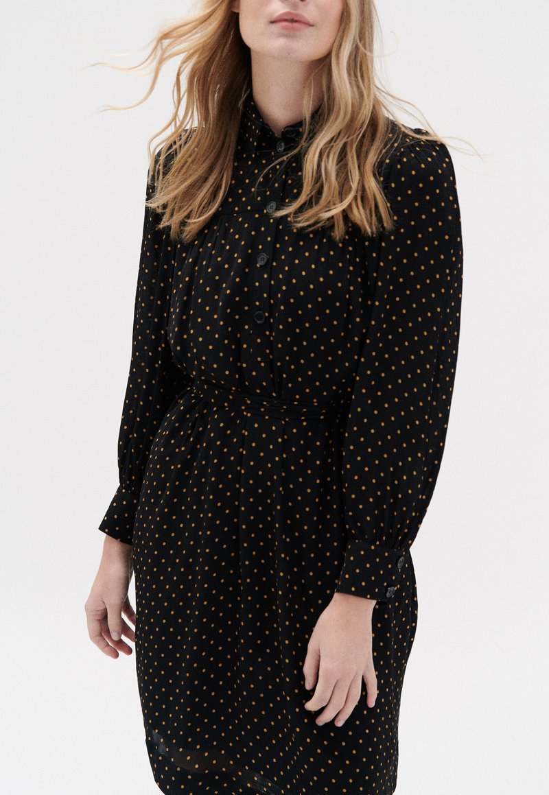 Zwarte jurk met lange mouwen en knoopmanchetten, gekleed met gele stippen en een riem op de taille, wat een getailleerde uitstraling geeft.