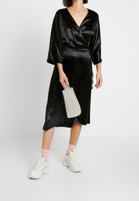 Musta satiininen kietaisumekko, jossa on kolme neljännesosaa hihasta, vyötäröltä kireä ja sivusolmu. Yhdistetty suurikokoisiin valkoisiin tennareihin ja helmillä koristeltuun laukkuun.