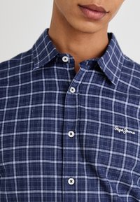 Camisa de cuadros azul marino hecha de algodón, con cuello abotonado, botones blancos y un pequeño logo bordado en el pecho.