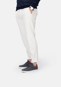 Zapatillas de tela azul marino con detalles de cuero marrón, combinadas con pantalones blancos de corte entallado. El modelo está de pie con las manos en los bolsillos, con un enfoque en el calzado.