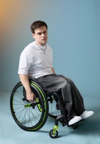 Wózek inwalidzki z czarną ramą i zielonymi akcentami. Użytkownik ubrany w białą koszulę i szare spodnie. Gładkie tło w jednolitym niebieskim gradiencie.