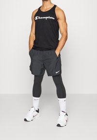 Camiseta de tirantes negra con el logo de "Champion", pantalones cortos grises, leggings negros, calcetines blancos y zapatillas deportivas con acentos negros. Fondo simple.