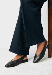 Fekete slingback cipők hegyes orral, puha bőr anyagból, varrott részletekkel és alacsony blokk sarkú, sötét nadrággal párosítva.