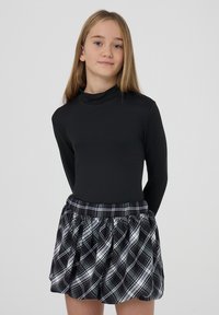 Ragazzina con capelli biondi lisci che indossa un top nero a maniche lunghe e una gonna a quadri bianco e nera, in piedi davanti a uno sfondo semplice.