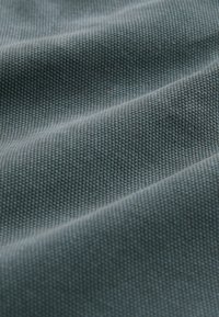Texturovaná látka v jemné šedomodré barvě, vykazující jemný tkaný vzor s ladnými záhyby a stíny, které zvýrazňují její strukturu.