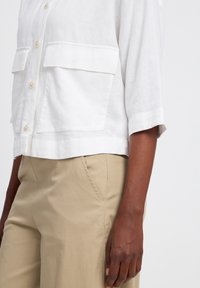 Witte linnen shirt met korte mouwen, grote borstzakken en parelknopen, gecombineerd met beige broek. Minimalistisch ontwerp met subtiele branding.