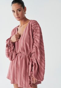 Modell mit langen Ärmeln in einem strukturierten rosa Kleid, stehend vor einem hellen Hintergrund und die Hand nahe ihrer Brust haltend.
