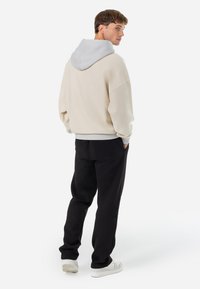 Lichtgekleurde oversized hoodie met een grijze capuchon, gecombineerd met zwarte gebreide broeken en witte schoenen, gezien van achteren.