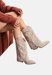 Bottes de cheville en suédine faux beige clair avec des bouts pointus, des accents cloutés et un talon bloc de hauteur intermédiaire, présentant un détail de boucle sur le côté.