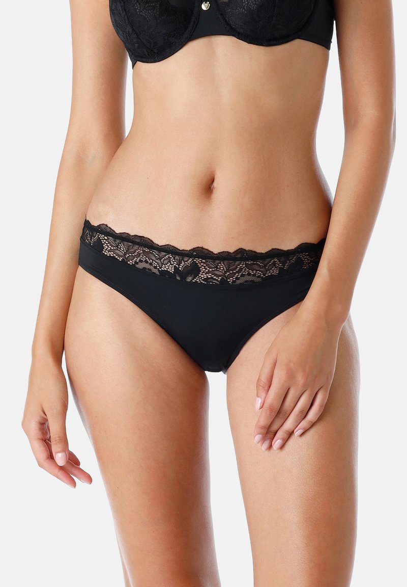 Donna che indossa lingerie nera con pizzo e un reggiseno abbinato, in piedi contro uno sfondo neutro con una mano appoggiata sulla coscia.