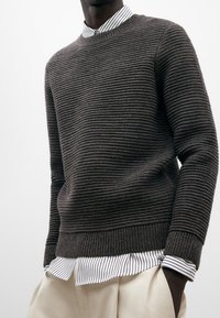 Suéter de punto texturizado en gris oscuro con rayas horizontales, cuello redondo y puños acanalados, superpuesto a una camisa de rayas blanca y negra.