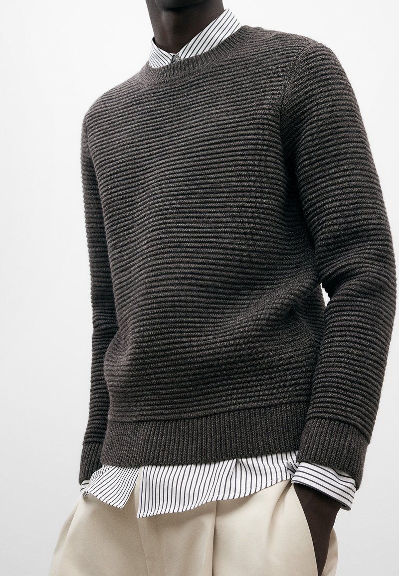 Suéter de punto texturizado en gris oscuro con rayas horizontales, cuello redondo y puños acanalados, superpuesto a una camisa de rayas blanca y negra.