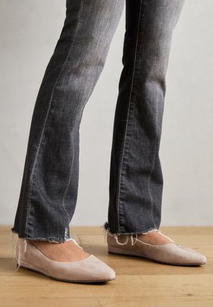 Světlé béžové semišové baleríny se špičatou špičkou, nošené s roztrhanými černými džínami s roztřepenými lemy. Pozadí z přírodního dřevěného podlahy.