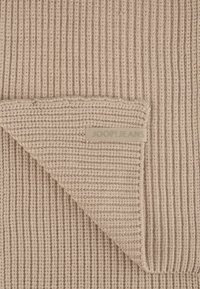 Couverture en tricot beige avec une texture côtelée et un coin replié, ornée d'un label "JOOP! JEANS" en marron clair cousu sur le bord.